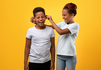 Fototapeta premium Siblings fights, quarrels, relationships concept. Naughty older sister pulling her little brother ears, yellow studio background. African american children having fight, teen sister teasing brother