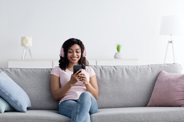 Fototapeta premium Lady listening music and chatting on phone, enjoy relax on couch at home. Satisfied millennial pretty black female in wireless headphones typing on smartphone on couch alone in living room interior