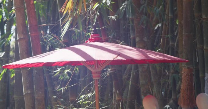 Red paper umbrella used as decoration at a local market in Uthai Thani, Thailand, placed among bamboo trees with natural sunlight and soft shadows creating a calm outdoor atmosphere.