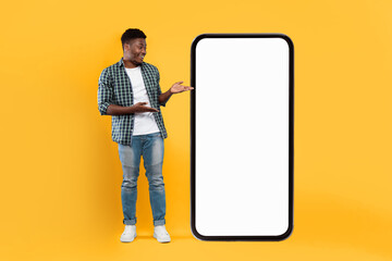 A man stands next to a large blank smartphone in a studio with a yellow background. He is smiling and gesturing towards the device. The setting is bright and eye-catching.
