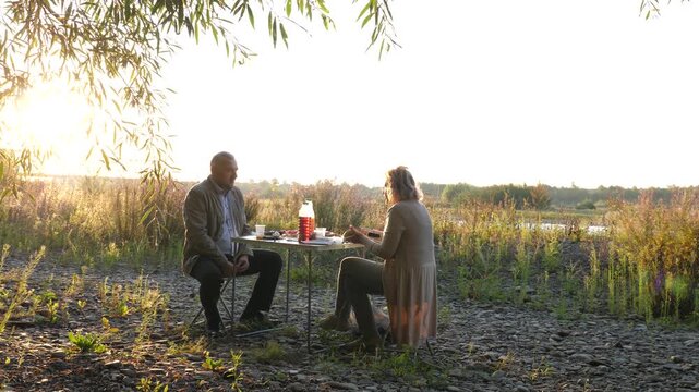 A couple sits at a table on an open riverbank talking and eating during a warm long outdoor rest