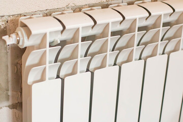 A white radiator mounted on a wall. The radiator has a modern design with vertical fins. The background shows a bare wall with exposed bricks.