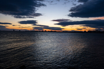 a dramatic sunset over a calm body of water, with dark clouds hovering above a distant city skyline silhouetted against the glowing orange horizon,  Sunny beach, Bulgaria
