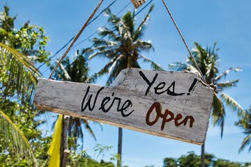 Fototapeta premium A rustic wooden sign reading “Yes We Are Open” hangs in front of lush tropical palm trees, creating a welcoming and relaxed island atmosphere. The image conveys hospitality, tourism, small business,