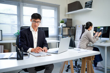 Fototapeta premium Asian business man sitting at desk, using laptop computer in open space office, female colleague is sitting and working in the back