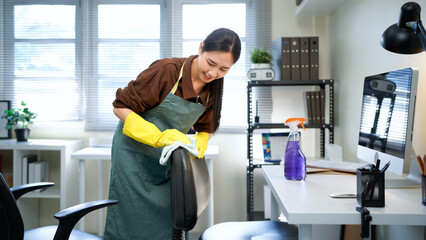 Young asian woman, daily janitor service lady in uniform with antiseptic spray bottle and wet cloth rag cleaning chair and table surfaces in modern office room interior