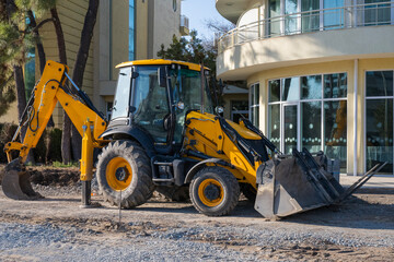 a yellow backhoe loader parked on a dirt path in front of a modern beige residential building with large glass windows.