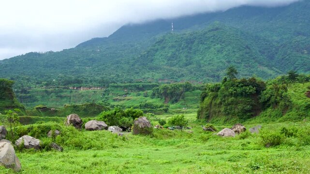 Scenic panning shot of lush green grassland with scattered large boulders and a misty mountain backdrop in Ranu Manduro, Indonesia. Beautiful nature landscape of East Java with rocky terrain and cloud