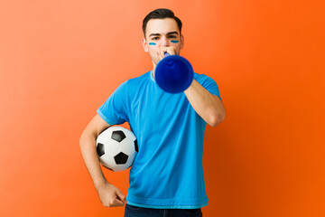 Soccer fan holding a ball and blowing a cheering horn, celebrating and supporting his favorite...