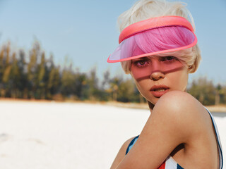 A close up portrait of a young woman on a sunlit beach, wearing a pink visor and swimsuit,...