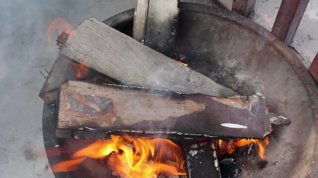 An elderly woman warms herself by adjusting firewood in an open fire, showing survival, warmth, and everyday rural life during harsh winter conditions in an Indian village.