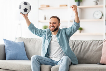 Excited young guy watching football on TV, lifting ball above his head, celebrating goal or victory...