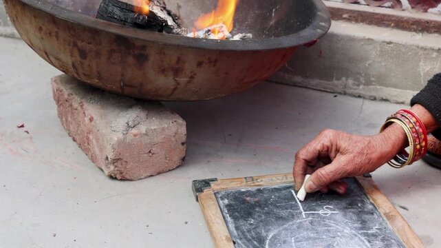 An elderly woman writes on a slate with chalk while a wood fire burns nearby for warmth, showing lifelong learning, resilience, and rural winter life in India.