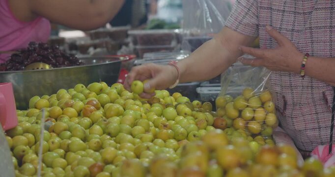 Fresh jujube fruits displayed in baskets while a customer selects and puts them into plastic bags at a local Thai market, showing everyday market life and self-service fruit shopping.