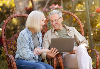 Happy elderly couple relaxing in wicker chairs in open air and using laptop computer, surfing...
