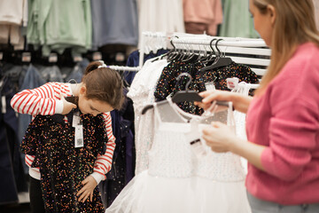A mother and her young daughter shopping together choose sequined dresses on hangers in a brightly...