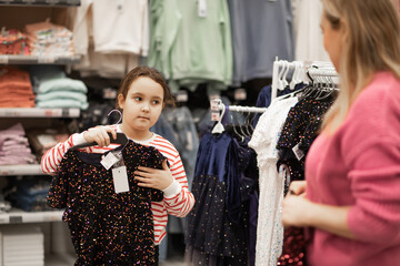 A mother and her young daughter shopping together choose sequined dresses on hangers in a brightly...