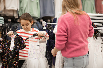 A mother and her young daughter shopping together choose sequined dresses on hangers in a brightly...