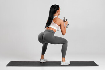 Athletic girl doing exercise for glutes with with dumbbells on gray background. Fitness woman working out
