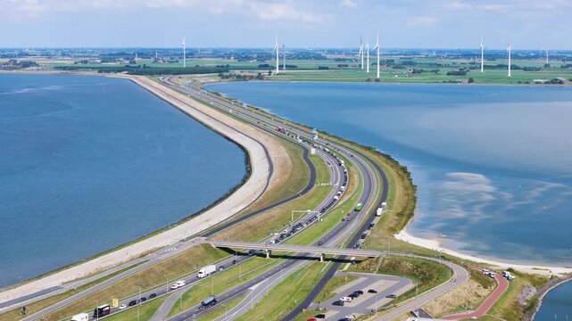 heavy traffic flows alongside blue sea curving highway section tall wind turbines dot distant green coastline ahorizon afsluitdijk netherlands long horizon transportation 