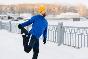 Young man stretching leg quadriceps in snowy park, warming up for winter running, staying fit and...