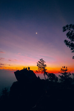 Silhouette of a group of people climbing on a large cliff. Colorful sky of orange, yellow, pink, and blue in the morning at sunrise. There are mountains and trees around with copy space on top.