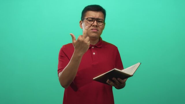 Young man wearing glasses and red polo holding book shows middle finger in green studio; frustration defiance.