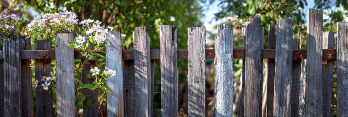 A fence made from recycled wooden planks and metal scraps encloses a suburban backyard. Green plants and flowers grow around the fence on a sunny day, banner
