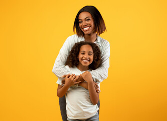 Mother's love. Happy loving african american mom cuddling her cute daughter from the back, standing over yellow background with free space and smiling to camera