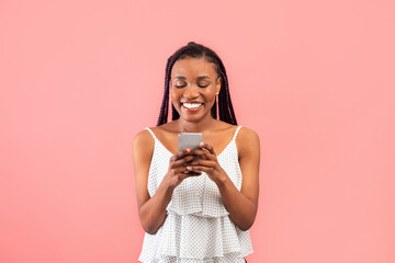 Happy young black woman using smartphone, chatting online, watching video on pink studio background. Lovely African American lady surfing web, sending sms, posting in social media