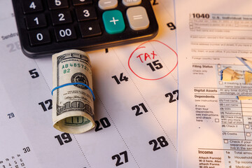 April 15 Tax Day deadline with red circle on a desk calendar near color IRS Form 1040 and a hundred dollar bill in sunny day light.