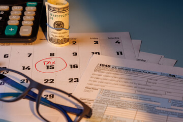 Tax Day deadline with a red circle on 15 and IRS Form 1040 in color near blue glasses and a calculator in bright morning sunlight today.