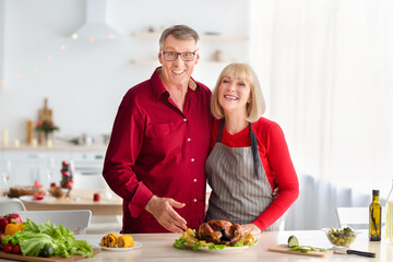 Cheerful senior couple posing and smiling at camera, cooking turkey for Christmas or Thanksgiving dinner, standing at table, preparing traditional holiday meal in kitchen