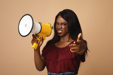 Emotional attractive african american millennial lady with loudspeaker pointing at camera over beige studio background, angry pretty black woman with megaphone getting attention