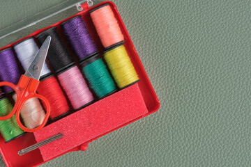 Spools of colorful sewing thread, scissors and needles in red plastic box on green background.