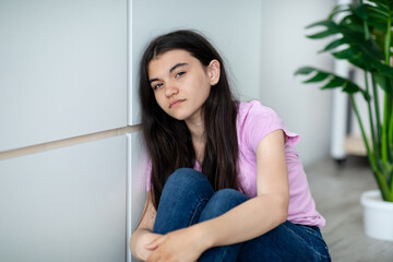 Portrait of Indian teen girl feeling upset and depressed, looking at camera, sitting near wall at...