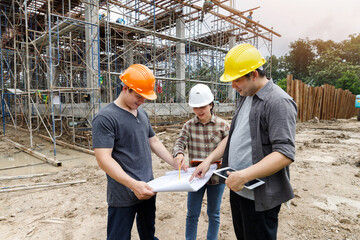 Group of Asian engineers and foreman holding blueprint discussing project at building site. Civil engineering team inspecting structure and scaffolding using architectural plans outdoors.