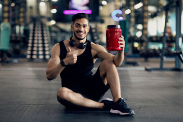 Obraz premium Handsome Muscular Middle Eastern Guy Advertising Fitness Supplements While Posing At Gym, Young Arab Male Athlete Holding Red Container With Whey Protein Powder And Showing Thumb Up At Camera