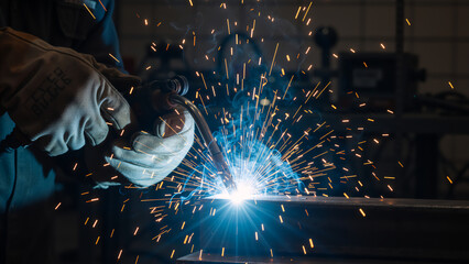 Close-up of a welder's gloved hands performing welding. Bright sparks and blue arc light during metal fabrication in an industrial workshop