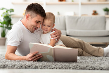 Naklejka premium Paternity Leave Leisure. Caring Young Dad Reading Book To His Adorable Infant Son While Relaxing Together On Floor In Living Room, Happy Father Bonding With Toddler Baby Kid At Home, Closeup