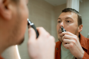 Man is using an electric trimmer to remove nose hair while looking at his reflection in a bathroom mirror, practicing daily self-care and personal grooming for a clean appearance