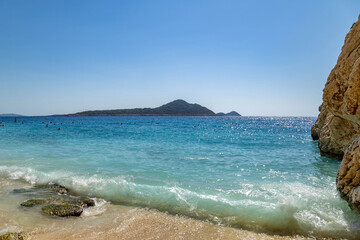 Obraz premium Turquoise sea waves crashing on sandy Kaputas Beach with limestone cliffs and island in the distance. Famous scenic coastline of the Mediterranean Sea. September, Kas-Kalkan region, Turkey.