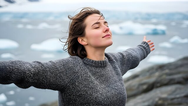 Young woman standing with arms outstretched on rocky coast in front of icebergs, breathing fresh air and enjoying freedom in cold nature of Greenland, travel and mindfulness concept