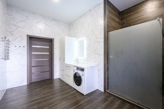 modern bathroom with marble-patterned white walls, dark wood-effect flooring, built-in white cabinetry, washing machine, frosted glass shower screen, and heated towel rail
