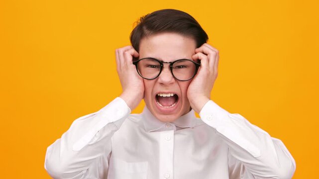 A young boy with glasses displays a frustrated expression while holding his head with both hands. The background is bright orange, creating a bold contrast. He appears to be in a playful situation.