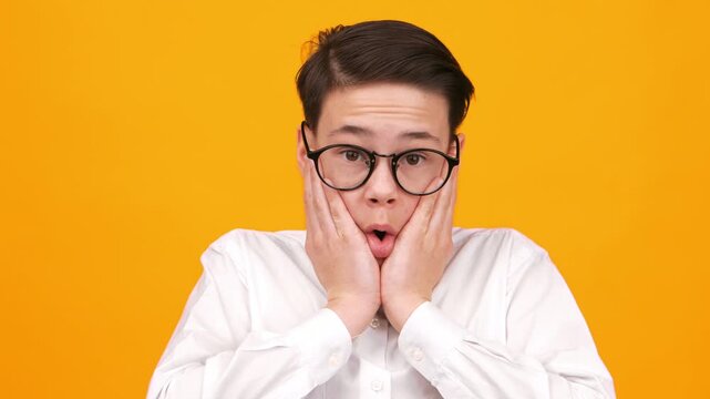 A boy wearing glasses stands with his hands on his face. He expresses surprise and shock as he looks directly at the camera. The background is a bright orange color.