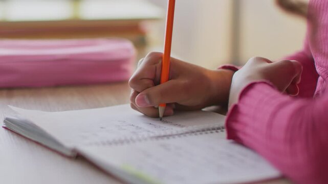 A person writes notes in a notebook with a bright orange pencil while sitting at a table. There are school supplies around in a sunny room. The focus is on the hand and writing action.