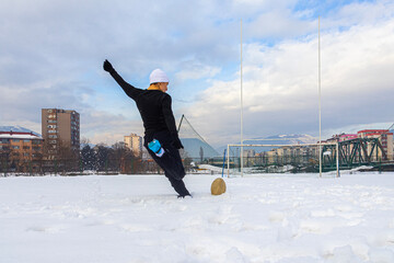 Athletic man kicking a rugby ball on a snowy urban sports field, seen from behind during winter training, with goal posts, city buildings and mountains creating a dynamic outdoor scene.