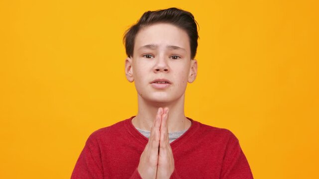 A boy stands with hands together as if requesting something. He faces the camera directly against a bright yellow background while recording a video. His expression suggests urgency.