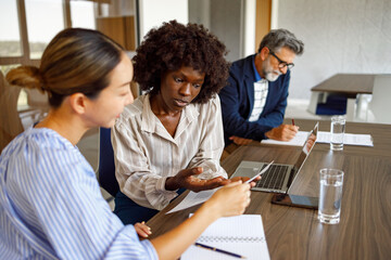 Focused female professionals reading documents while sitting beside male colleague in modern boardroom during business meeting